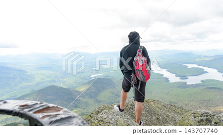 Hiker with red backpack standing on rocky summit above expansive green mountain valley and winding lakes in a scenic wilderness landscape 143707740