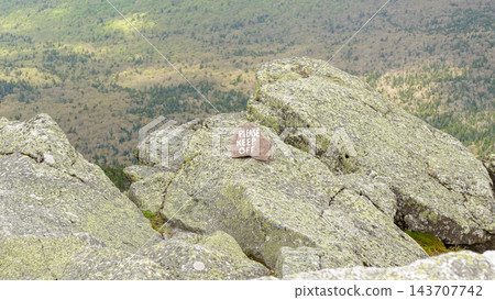 Weathered please keep off sign on lichen covered granite rocks overlooking a forested mountain landscape from a rugged summit Weathered please keep off sign on lichen covered granite rocks overlooking a forested mountain landscape from a rugged summit 143707742