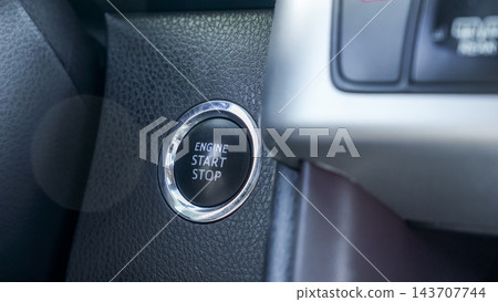 Close up of illuminated engine start stop button with chrome trim beside dark textured dashboard in a modern vehicle interior Close up of illuminated engine start stop button with chrome trim beside dark textured dashboard in a modern vehicle interior 143707744