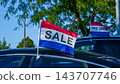 Colorful sale flags attached to parked vehicles beneath leafy trees at an outdoor pre owned automobile lot 143707746