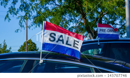 Colorful sale flags attached to parked vehicles beneath leafy trees at an outdoor pre owned automobile lot Colorful sale flags attached to parked vehicles beneath leafy trees at an outdoor pre owned automobile lot 143707746