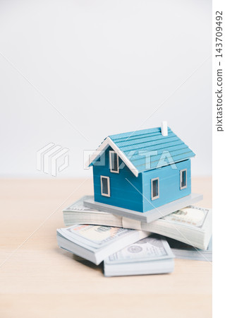 A blue model house sits atop stacked banknotes in a vertical composition with generous light copy space above, illustrating a home savings goal, purchase planning, and housing costs. A blue model house sits atop stacked banknotes in a vertical composition with generous light copy space above, illustrating a home savings goal, purchase planning, and housing costs. 143709492
