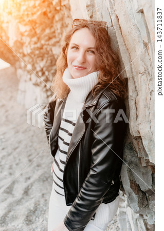 Woman smiling portrait leaning against a stone wall while wearing a leather jacket and striped sweater in golden light 143711837