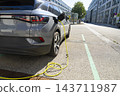 A grey electric car is plugged into a charging station on a street. 143711987