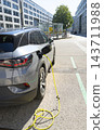 A grey electric car is plugged into a charging station on a street. 143711988