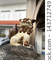 A white Shisa sits with a row of red-tiled houses in the background. 143712749