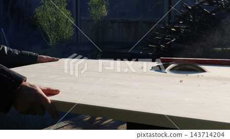 Off-camera, a carpenter carefully guides a wide beech wood panel toward the rotating saw blade of a machine. The process of rip-cutting timber takes place outdoors on a sunny day. Off-camera, a carpenter carefully guides a wide beech wood panel toward the rotating saw blade of a machine. The process of rip-cutting timber takes place outdoors on a sunny day. 143714204
