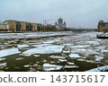 Winter Cityscape With Frozen River, Ice Floes and Urban Skyline Under Overcast Sky With Construction Cranes 143715617