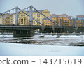 Winter Cityscape With Frozen River and Steel Suspension Bridge Spanning Historic Riverfront Buildings 143715619