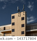 Sunlit Beige Apartment Building Facade With Square Windows, Balconies And Antenna Against Blue Sky 143717804