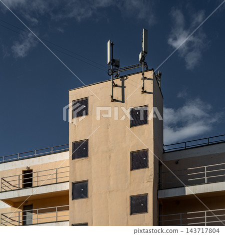 Sunlit Beige Apartment Building Facade With Square Windows, Balconies And Antenna Against Blue Sky 143717804