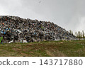 Household waste is piled across a dump beside a grass field, with trees in the background under an overcast sky 143717880