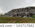 A mound of mixed waste fills a landfill beside a grassy field, with trees in the distance under an overcast sky during the day 143717882