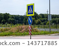 A pedestrian crossing sign and a blue direction sign stand beside a road near a fence and trees during the day 143717885
