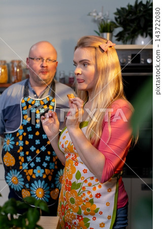 Blonde white woman tasting batter spoon, smiling while bald white man watches nearby in cozy kitchen with jars, plants, floral aprons, wooden counter and basil pot Blonde white woman tasting batter spoon, smiling while bald white man watches nearby in cozy kitchen with jars, plants, floral aprons, wooden counter and basil pot 143722080