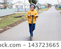 A joyful boy wearing glasses and a helmet rides a scooter along a residential street in autumn. 143723665