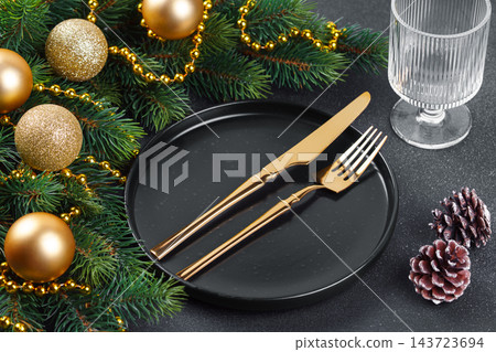 Elegant Christmas table setting with gold cutlery on black plate and decorated spruce branches 143723694
