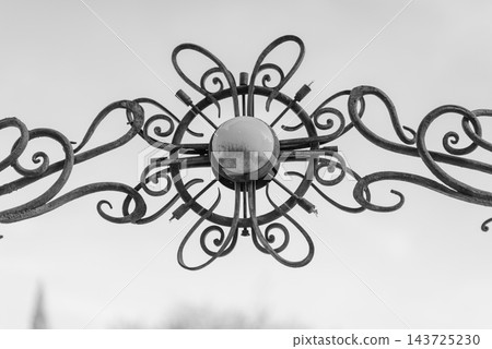 A detailed close-up view of an ornate metal gate with circular design featuring eight flowing arms and a central light fixture or reflective surface A detailed close-up view of an ornate metal gate with circular design featuring eight flowing arms and a central light fixture or reflective surface 143725230