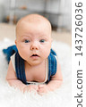 Baby 5-6 months old in blue overalls lies on tummy on a white blanket at home, exercising muscles and looking at camera 143726036