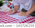 A person writes on a sheet of paper at a red and white checkered table outdoors during the day 143726159