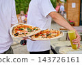 Two workers hold pizzas beside a drink at an outdoor food stall in daylight, with customers and trees in the background 143726161