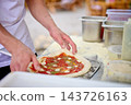 A baker arranges toppings on pizza dough at a kitchen workbench, with ingredient containers nearby during food preparation 143726163