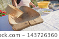 Old wooden dough trough with round wooden sieve on white patterned tablecloth outdoors under warm daylight lighting filling foreground with baking bowls and utensils in background 143726360