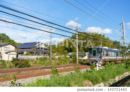 一列 225 系列通勤列車沿著寧靜的鐵路線行駛,秋日的天空晴朗,遠處是綿延的村莊。 一列 225 系列通勤列車沿著寧靜的鐵路線行駛,秋日的天空晴朗,遠處是綿延的村莊。 143731443