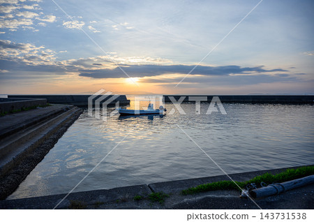 一艘藍色的小漁船漂浮在平靜的海面上,夕陽西下,在混凝土防波堤環繞的漁港中,營造出一幅寧靜的傍晚景象。 一艘藍色的小漁船漂浮在平靜的海面上,夕陽西下,在混凝土防波堤環繞的漁港中,營造出一幅寧靜的傍晚景象。 143731538
