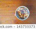 A takeout container of thinly sliced spicy meat and onions over rice, sprinkled with sesame seeds, on a wooden table. Top-down view of a packaged meal for delivery or carry-out. 143733373