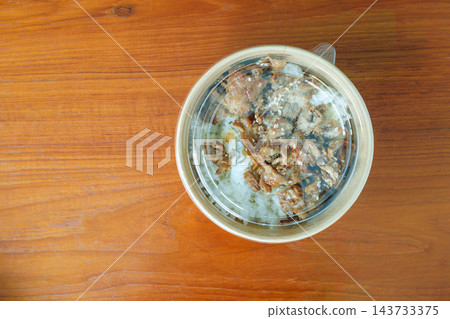 Grilled meat and rice topped with sesame seeds in a round takeout container on a wooden table. High angle view of a convenient packaged meal. Grilled meat and rice topped with sesame seeds in a round takeout container on a wooden table. High angle view of a convenient packaged meal. 143733375