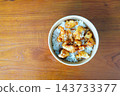 A bowl of crispy fried chicken pieces with sesame seeds served over white rice in a disposable container on a wooden table. High-angle view of a take-out style Asian meal. 143733377