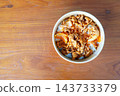 High-angle view of a bowl of spicy stir-fried pork with onions and sesame seeds served over white rice in a paper take-out container on a wooden table. 143733379