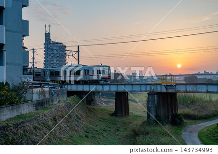 城市景觀描繪了一列銀色的火車駛過鐵路橋，橋上架著電線，橋墩和綠草茵的河岸，背景是淡橙色的天空和低垂的太陽。 143734993