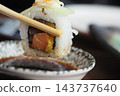 Chopsticks hold a sushi roll above a serving dish during a meal 143737640