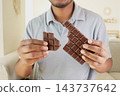 A man is holding two pieces of a chocolate bar while standing in a room 143737642