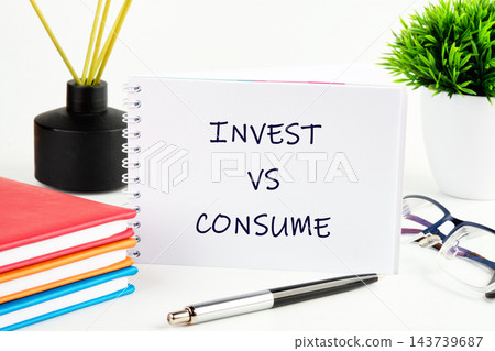 A notebook reads invest versus consume beside stacked notebooks, a pen, glasses, and a plant on a desk in a bright indoor setting 143739687
