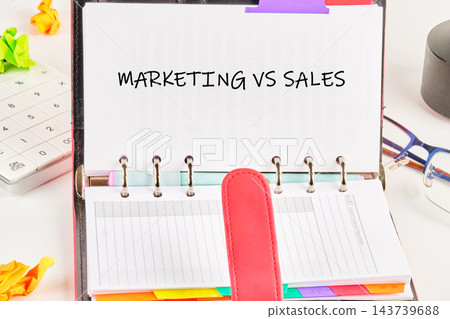 Marketing versus sales is written in an open planner on a desk with a calendar and office supplies during daytime Marketing versus sales is written in an open planner on a desk with a calendar and office supplies during daytime 143739688