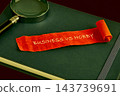 A red ribbon marked business vs hobby rests on a green book beside a magnifying glass on a tabletop, with no time or place shown 143739691