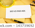 A paper sheet labeled SEO vs paid ads lies on a desk with a pen, glasses, and magnifying glass during a marketing work session 143739692