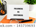 Notebook with the words followers versus customers sits on an orange folder beside a magnifying glass and pen on an office desk in daylight 143739693