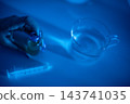 Hands pouring liquid from dropper into glass measuring cup under cool blue clinical lighting with syringe on sanitized surface, close-up precision dosing scene for laboratory or medical use 143741035