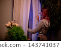 Woman with red hair reads outdoors. Caucasian woman with red hair looking through window at dusk. Quiet evening scene with woman in cozy apartment reading on her phone 143741037