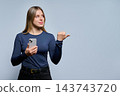 Smiling young woman holding smartphone pointing thumb aside studio 143743720