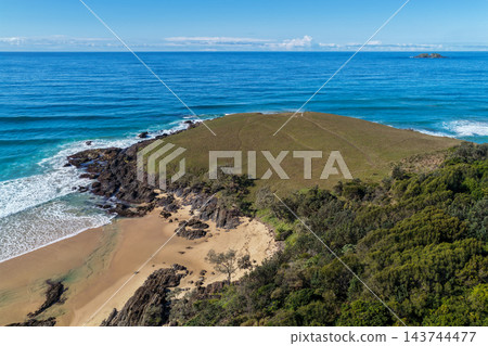 Aerial view across a grassy headland at Sapphire Beach, with rocky shoreline, breaking surf, a sandy cove and a distant offshore island near Coffs Harbour. 143744477
