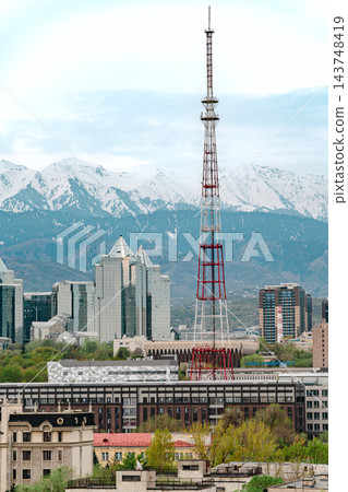 Tall red and white television tower rising above Almaty city buildings with snowy mountains behind 143748419