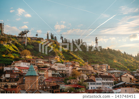Cable car cabins over old town houses with Mother of Georgia statue on the hill at sunset in Tbilisi 143748451
