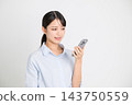 一位微笑的女上班族身穿淺藍色襯衫，在白色背景下操作智慧型手機螢幕。 143750559