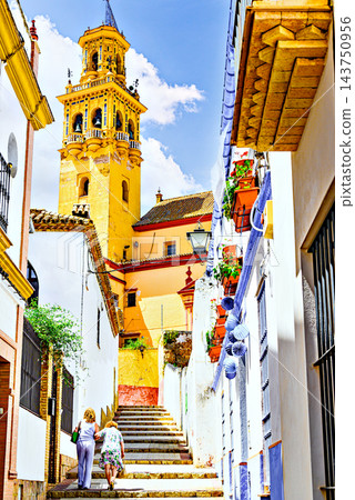 Two women walk up the street leading to the Church of Santiago in Alcala de Guadaira, with the church's bell tower in the background; Seville. Two women walk up the street leading to the Church of Santiago in Alcala de Guadaira, with the church's bell tower in the background; Seville. 143750956