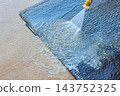 Pressure washer nozzle sprays water over dark car carpet surface outdoors, rinsing dirt toward edge 143752325
